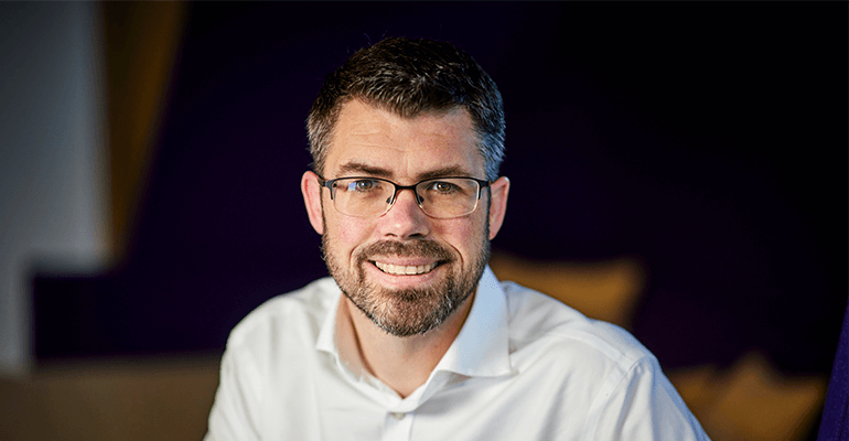 A middle-aged man with short grey hair and a beard with glasses smiling.