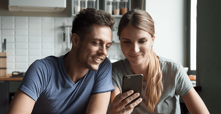 A blonde and brown haired couple both smiling at a phone screen together.