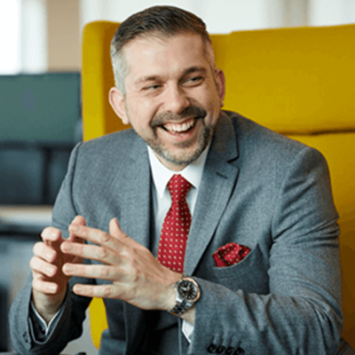 A man with short grey hair and a beard smiling away from the camera sat on a yellow chair in a grey suit.