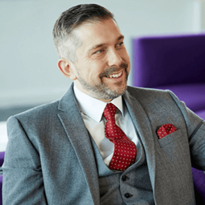 A man with short grey hair and a beard smiling wearing a grey suit.