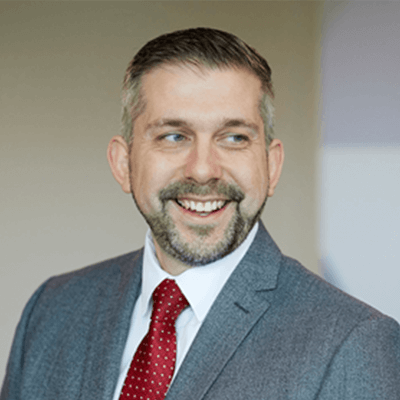 A man with short grey hair and a beard smiling wearing a grey suit and red tie.