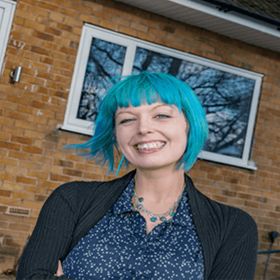 A woman with a short blue bob smiling with a blazer and blouse on in front of a house.