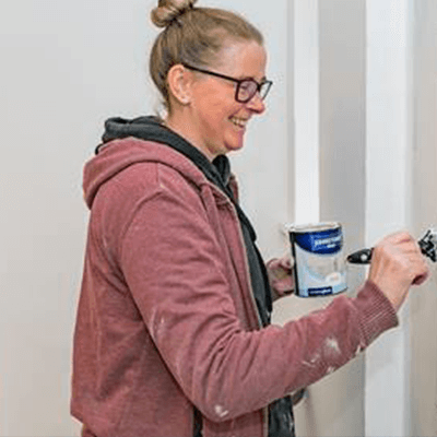 A woman with blonde hair in a bun smiling whilst painting a wall white.