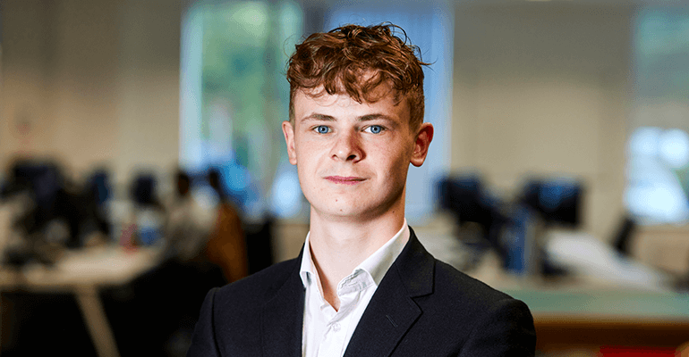 A young man with ginger curly hair smiling wearing a back suit and white shirt.