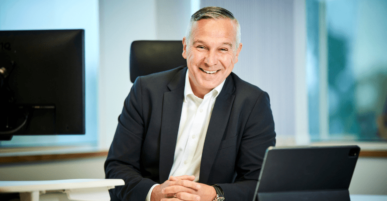 Chris Baguley, smart professional man in a suit without a tie, casually sat at an office desk smiling.