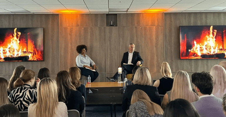 Christina Dejonge, a Caribbean woman, and Chris Baguley, a white male in a suit, are sat on a stage, talking in front or an audience.