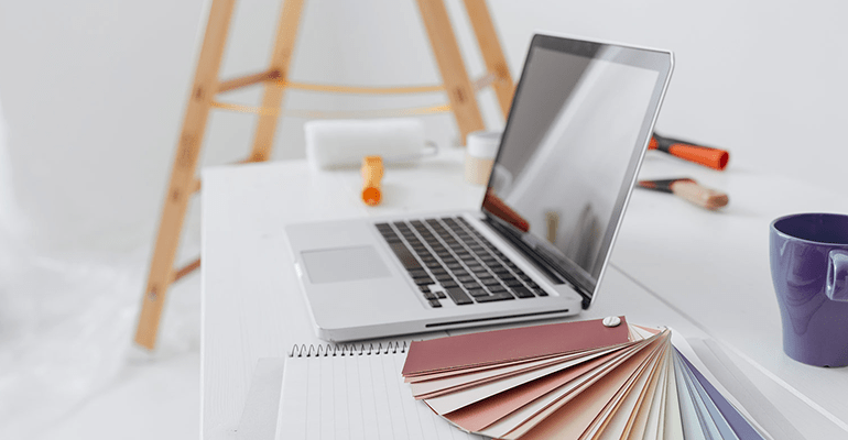 laptop on a desk next to a notepad and colour swatch with a ladder behind - l