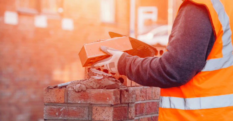 Male builder in orange vest laying a brick on a wall.