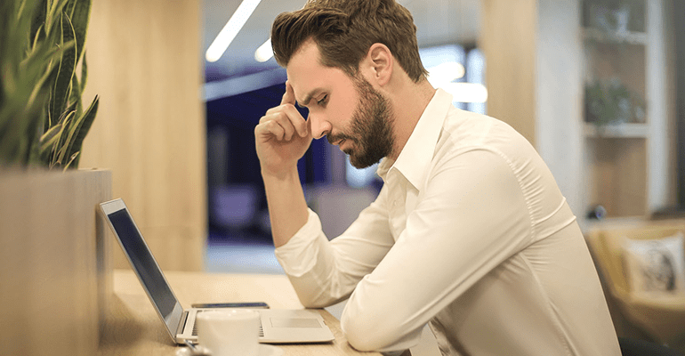 man sat at a desk in front of a laptop with his head in his hand - l