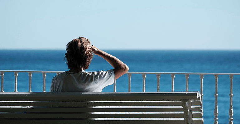 man sat on a white wooden bench looking out to the sea - l