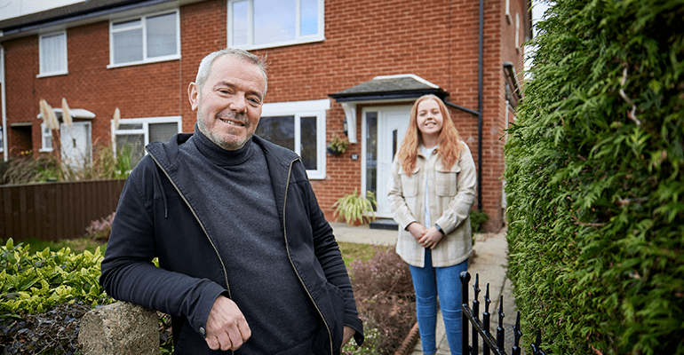 man with grey hair stood in front of a garden gate next to a woman in front of a house - l