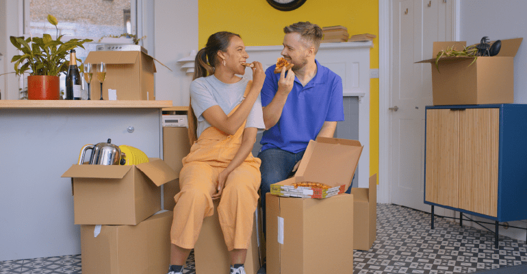 Young couple eating pizza on cardboard packing boxes.