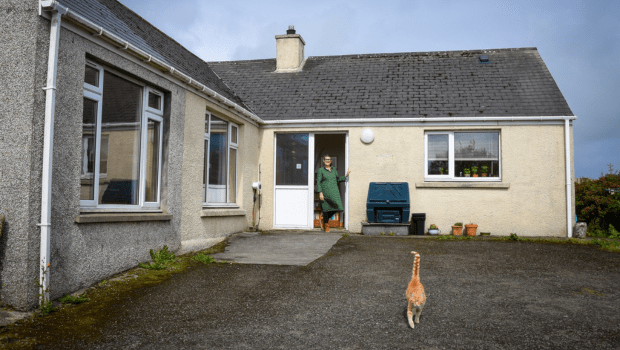 Older woman standing in the doorway of a bungalow as a ginger cat walks towards the camera.