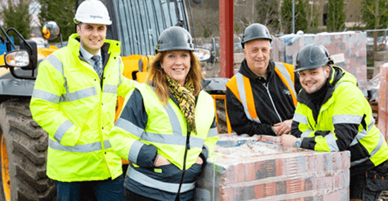 group of people wearing high vis jackets and hard hats stood by a pallet of bricks - l