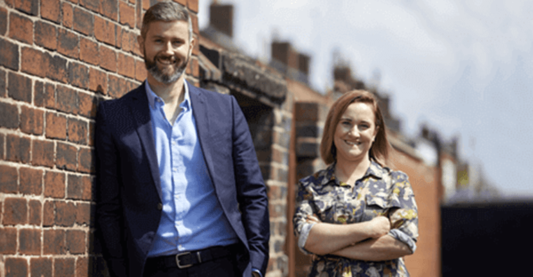 man in a blue suit stood with a woman smiling at the camera next to a red brick wall - l