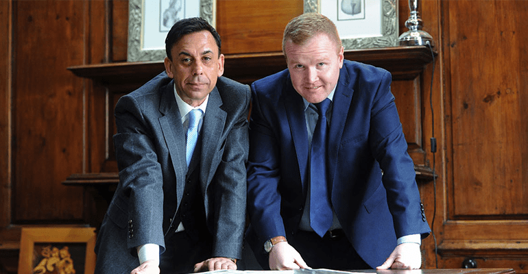 two men in suites leaning over paperwork on a table in a wooden panelled room - l