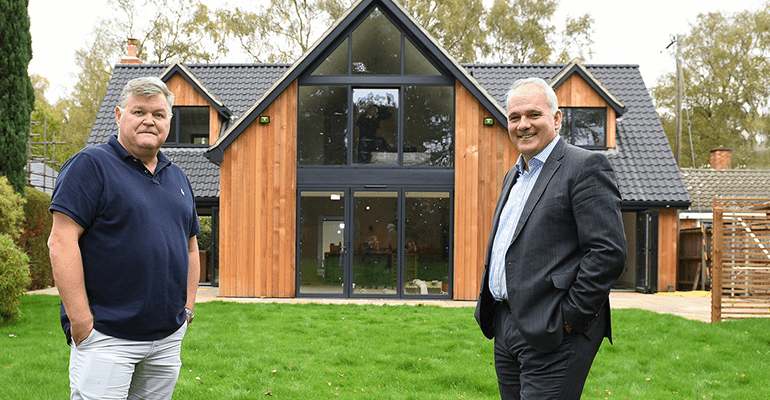 Two men stood apart smiling with a two story house in the background - l