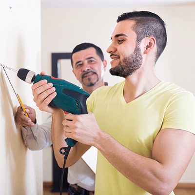 An older man looking at a younger man drilling into a wall.