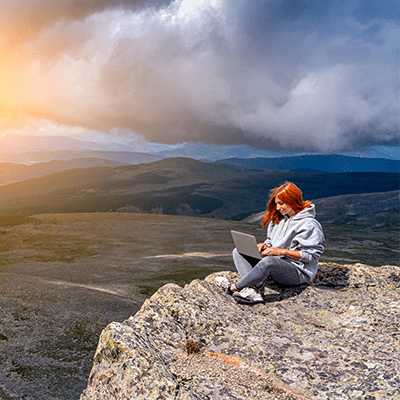 red haired woman sat on a cliff working on a laptop with mountains in the background