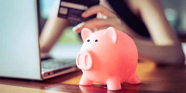 A pink piggy bank with a blurred woman in the background holding a card and looking at a computer.