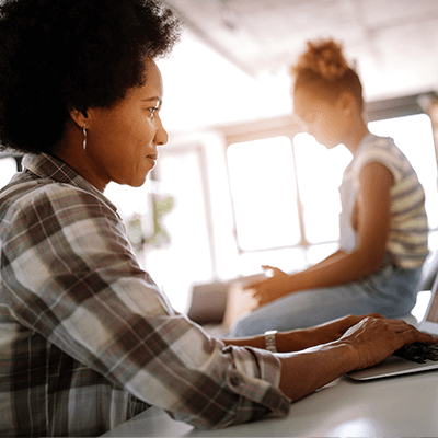 An African American mother working on her laptop while her daughter sits on the counter top.