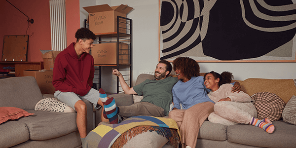 A wide shot of a family talking on a couch with moving boxed around them.