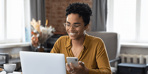 A woman sat down smiling at her laptop with a phone in her hand.