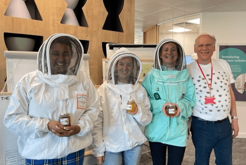 Four people stood together in bee suits holding jars of honey.