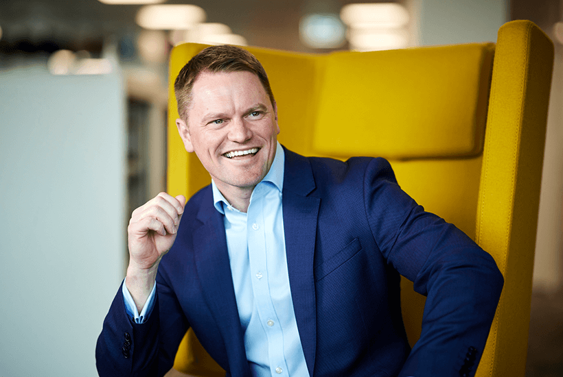 A man with short brown hair smiling wearing a navy blue suit sat on a yellow chair.