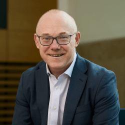 A professional male with a bald head and glasses smiling wearing a white shirt and grey blazer.