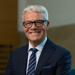 An older professional male with short grey hair and glasses smiling wearing a navy suit.