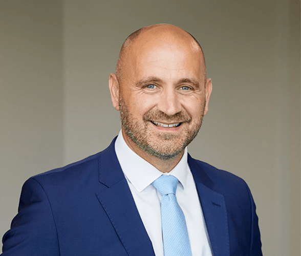 A bald man with a stubble smiling wearing a navy blue suit with a white shirt and a light blue tie.