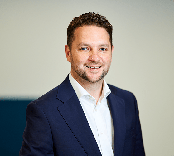 A man with short brown hair smiling wearing a navy blue blazer and white shirt.