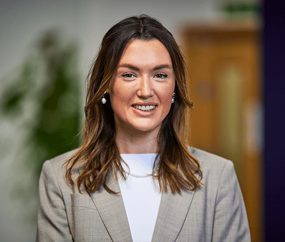 A young woman with medium length brown hair smiling with a beige blazer and white shirt on.