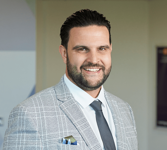 A man with short black hair and a stubble smiling wearing a grey suit.