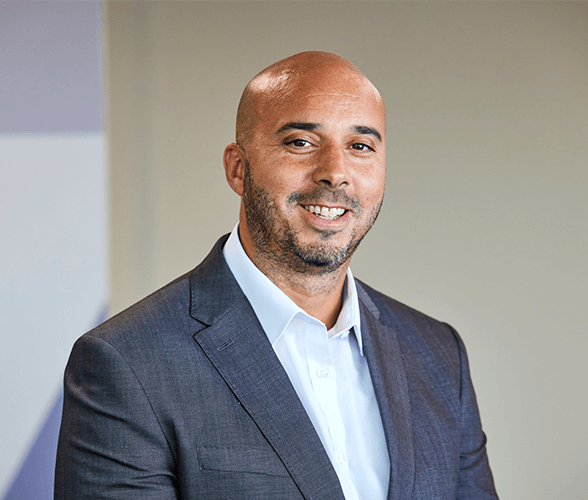 A bald man with a black beard smiling wearing a grey suit and white shirt.