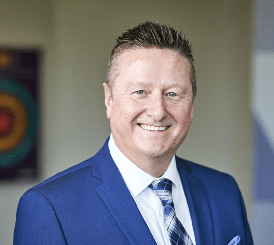 A man with short brown hair smiling wearing a grey suit.