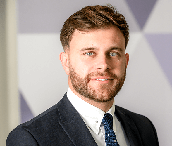 A man with short light brown hair smiling wearing a suit.