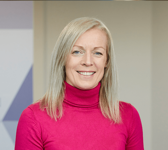 A woman with mid-length blonde hair smiling wearing a pink turtleneck.