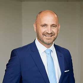 A bald man with a stubble smiling wearing a navy blue suit with a white shirt and a light blue tie.