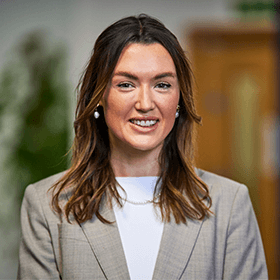 A young woman with medium length brown hair smiling with a beige blazer and white shirt on.