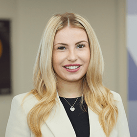 A woman with long blonde hair smiling wearing a beige blazer and black top.