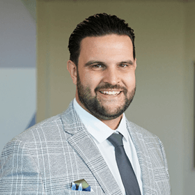 A man with black hair and a beard smiling in a grey suit.