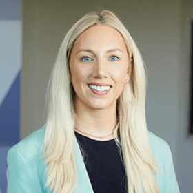 A woman with long blonde hair smiling wearing a black top with a blue blazer.