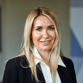 A woman with mid-length blonde hair smiling wearing a black blazer and white shirt.