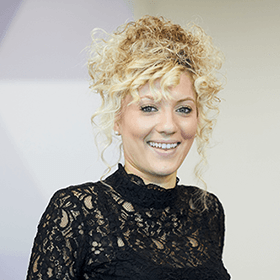 A woman with curly blonde hair in a ponytail smiling wearing a black lace dress.