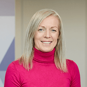 A woman with mid-length blonde hair smiling wearing a pink turtleneck.