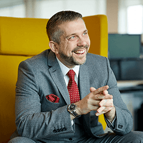 A man with short grey hair wearing a grey suit while smiling sat on a yellow chair.