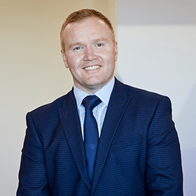 A man with ginger hair smiling wearing a navy blue suit.