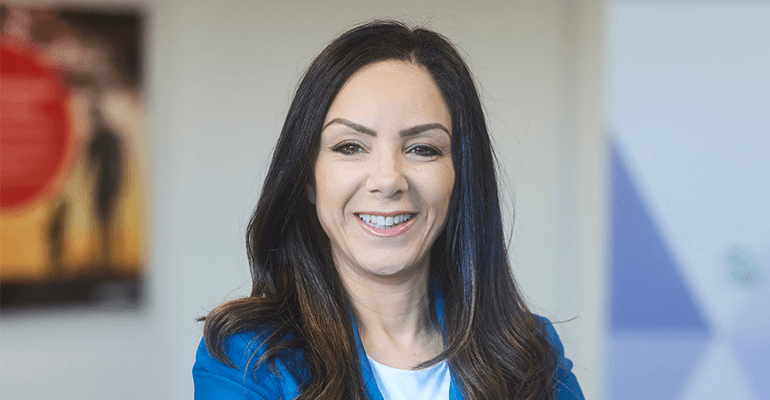 A woman with long brown hair smiling wearing a blue blazer with a white shirt.